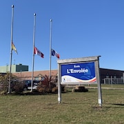 Un drapeau du Nouveau-Brunswick, un drapeau du Canada et un drapeau de l’Acadie sont en berne devant un établissement scolaire. Sur un panneau à l’entrée, il est écrit École L’Envolée.