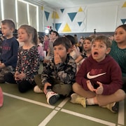 Des enfants d'une école primaire assis dans un gymnase, bouche bée devant les instruments de musique qu'ils ont reçus. 