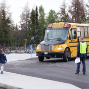 Une personne observe un parent avec son enfant marchant sur un trottoir, devant un autobus scolaire.
