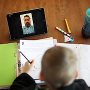 Un garçon photographié de haut devant ses cahiers d'école. Une tablette est posé devant lui et on y voit son professeur donnant le cours.