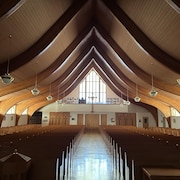 Allée centrale d'une église bordée de chaque côté de bancs en bois surplombée d'un plafond cathédrale en bois.