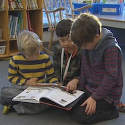 Trois élèves lisent un livre par terre dans une bibliothèque, devant une table et une étagère de livres.