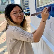 Emily Chu tient un pinceau dans sa main et est en train de peindre le mur où se trouvera la murale. 