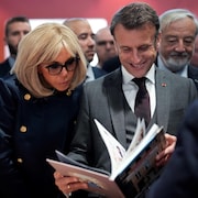 Emmanuel Macron, tout sourire, et sa femme Brigitte, concentrée, feuillettent un livre dans une foule.