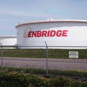 Un réservoir de pétrole de forme cylindrique entourée d'herbe au sol. En grosse lettre, c'est écrit sur le réservoir : Enbridge, le nom de l'entreprise.