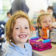 Une petite fille rousse assise à l'école devant sa boîte à lunch se tourne vers la caméra et sourit même s'il lui manque une des deux palettes en avant.