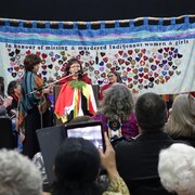 Une femme parle au microphone devant une broderie pour les femmes autochtones. 