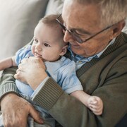Un homme âgé, assis sur un sofa, sourit en tenant un bébé dans ses bras.