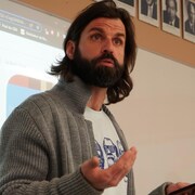 Un homme barbu enseigne devant une classe avec son téléphone à la main.