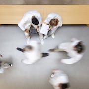 Des personnes en sarrau marchent dans un corridor; d'autres personnes sont assises et regardent une tablette et un cahier.