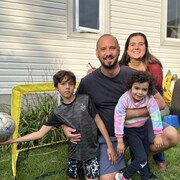 Portrait d'une famille devant le mur d'une maison. Une home souriant avec une barbe tiens une fillette de quatre ans dans un bras et un garçon de huit ans dans l'autre. Le garçon tient un ballon de soccer dans une main. La mère aux cheveux longs est accroupie derrière eux.