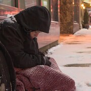 Une femme sans-abri assise sur un trottoir enneigé.