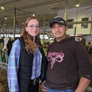 Une femme et un homme sourient à la caméra en posant dans une étable, quelques vaches en arrière-plan.