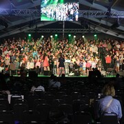 Deux responsables font face à des centaines de jeunes qui sont alignés sur une scène. Quelques personnes sont présentes dans la salle.