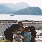 Une femme montre à un petit garçon comment regarder dans un  microscope. Le duo se trouve sur une plage au bord du fleuve.