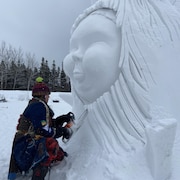 Une personne sculpte une visage en neige géant avec une scie mécanique.