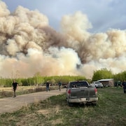 Des personnes et des véhicules sur une ferme à l'avant-plan. Au loin, de la fumée masque le ciel.