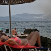 Des touristes prennent du soleil sur une plage en regardant la fumée qui s'élève d'une île voisine.