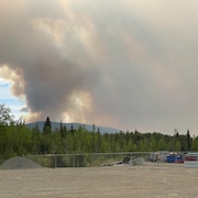 Une épaisse fumée s'élève au-dessus d'une montagne avec en avant-plan du matériel de construction gardé par une clôture.