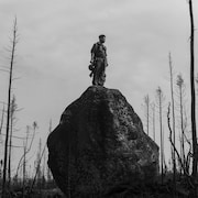 Photo en noir et blanc montrant un homme perché sur le haut d'une grosse roche, entouré d'arbres décimés par le feu. 