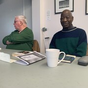 Deux hommes sont assis derrière une table sur laquelle se trouvent des livres et une tasse.