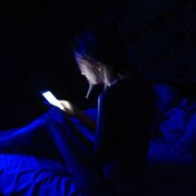 La jeune fille est assise sur un lit. Elle tient un téléphone mobile dans ses mains. Celui-ci éclaire la pièce sombre.