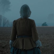 Une femme vêtue d'un costume d'époque est debout dans un champ à l'aube.
