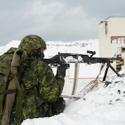 Un militaire tient un fusil mitraillette avec une ceinture de balles avec en arrière-plan une mine abandonnée.
