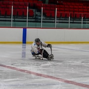François Alain est assis sur une luge et glisse sur une patinoire à l'aide de deux bâtons de hockey.