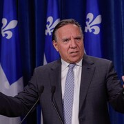 Francois Legault parle devant deux micros, les mains dans les airs. Derrière lui, il y a des drapeaux du Québec.