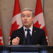 Le ministre Champagne en conférence de presse devant des drapeaux canadiens.