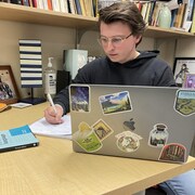 Un jeune homme est assis devant un ordinateur et prend des notes dans un cahier.