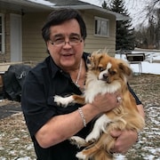 Frank Ostrowski est à l'extérieur de sa maison, il regarde la caméra et tient un petit chien entre ses bras. 