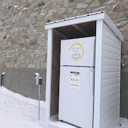 À l'extérieur, en hiver, un frigo est protégé par un abri, tout près du mur d'un bâtiment. 
