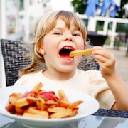 Une petite fille assise à l'extérieur devant une assiette de frites a la bouche grande ouverte et s'apprête à y faire entrer une frite.