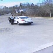 Debout près d'une auto de police dans un stationnement, un homme habillé en policier retire son blouson.