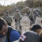 Des migrants marchent devant des soldats de la Garde nationale qui montent la garde derrière des barbelés.