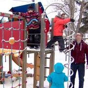 Des enfants jouent dans un module de parc l'hiver sous la surveillance de deux adultes. 