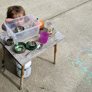 Une petite fille, cheveux aux épaules, joue à l'extérieur. Elle tient un bac transparent d'une main et s'appuie avec l'autre sur une table basse.
