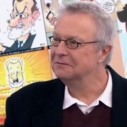 Michel Garneau souriant devant un écran où figure un collage de plusieurs de ses caricatures.