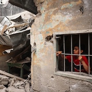 Un garçon se trouve derrière une fenêtre avec des barreaux dans une maison qui jouxte des décombres.