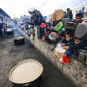 Une foule avec des contenants divers attend devant des marmites de nourriture.