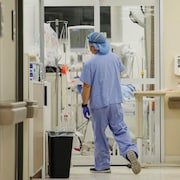 Un infirmier de dos et en uniforme marche vers une chambre d’une unité des soins intensifs. Il y a énormément d’équipement médical dans cette salle, qu’on voit au loin. L’homme est masqué et a aussi des gants. Il y a aussi une femme de dos, également en uniforme. 