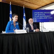 Les ministres Geneviève Guilbault et Jonatan Julien assis à la table d'une conférence de presse, à côté d'un écran où on peut lire : «Pour des projets de qualité, plus rapidement et à meilleur coût».