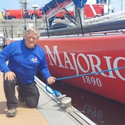 Georges Leblanc agenouillé à quai à côté d'un voilier à la coque rouge