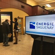 Une affiche d'Énergie Saguenay avec deux personnes en arrière-plan qui consultent des affiches. 