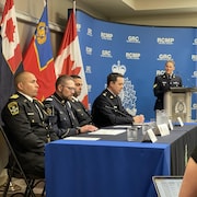 Quatre hommes en uniforme en conférence de presse assis à une table avec une femme, en uniforme, derrière un lutrin et un micro.