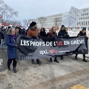 Des professeurs manifestent sur le campus de l'Université Laval. Ils portent une banderole et des pancartes. 