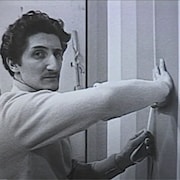 Guido Molinari colle du ruban adhésif sur une toile en regardant la caméra. 