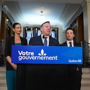 Geneviève Guilbault, François Legault, parlant au micro au lutrin, et Simon Jolin-Barrette devant un escalier à l'Assemblée nationale du Québec.